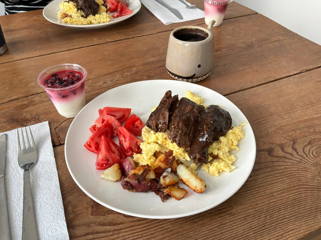 A plate featuring scrambled eggs topped with slices of beef, accompanied by diced potatoes and fresh tomato wedges. A small cup of yogurt with berries is visible in the background.