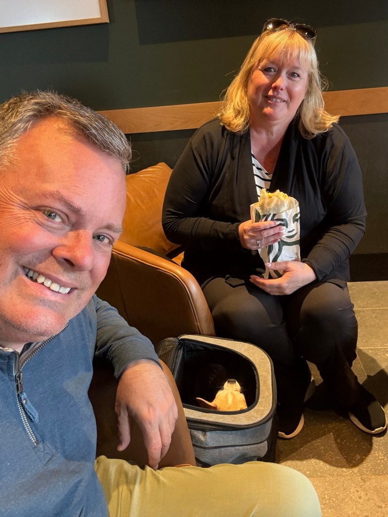 A smiling man taking a selfie with a woman sitting next to him in a café, holding food. The woman has blonde hair and is wearing a black cardigan, while the man is wearing a blue top.