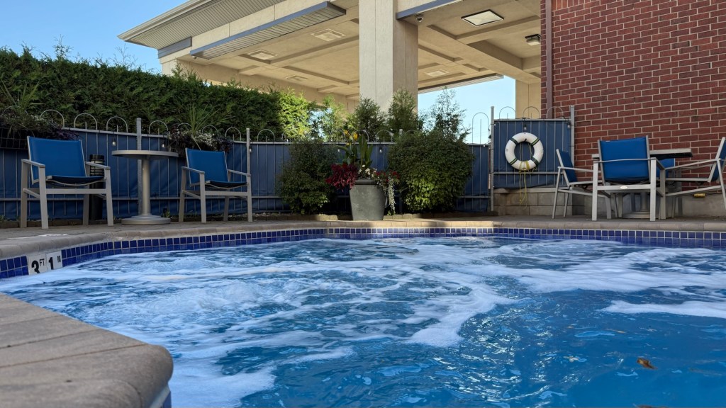 A hot tub with bubbling water, surrounded by blue chairs and a table. In the background, there are potted plants and a lifebuoy on a fence, with a building structure visible overhead.