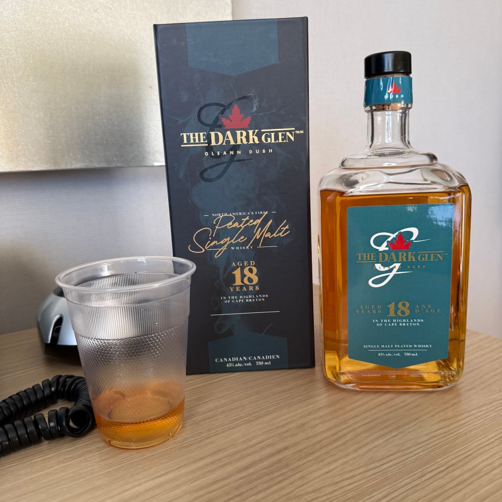 A bottle of The Dark Glen 18 Year Old Peated Single Malt Whisky next to a plastic cup containing whisky, on a wooden table. A telephone cord is visible in the background.