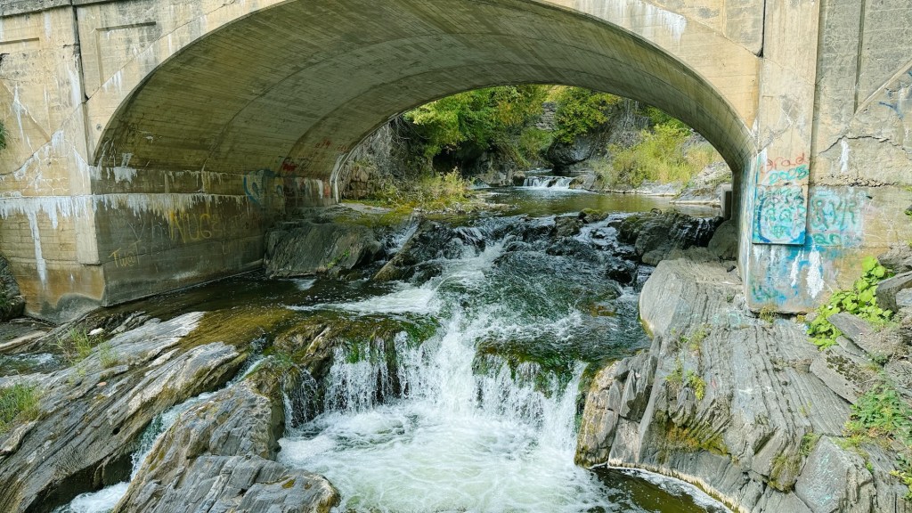 View of a rocky riverbed under a concrete bridge, with water cascading over rocks and greenery along the banks.
