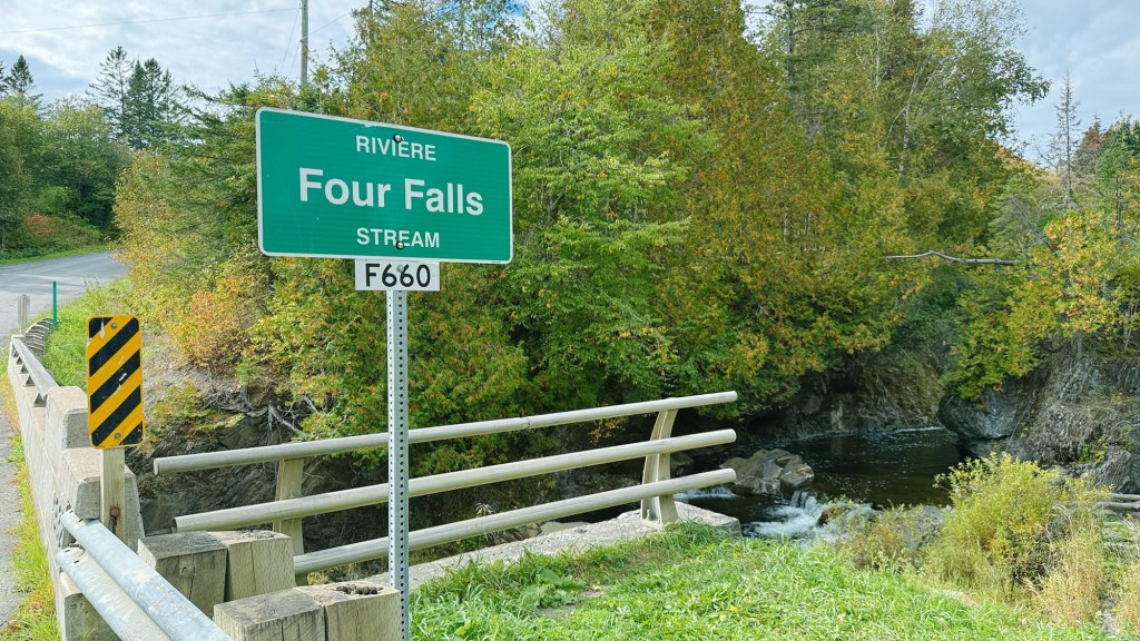 A green road sign indicating 'Four Falls Stream' next to a small bridge over a stream, surrounded by lush trees and vegetation.