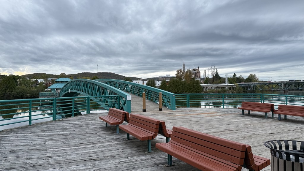 View of a wooden deck with benches overlooking a river, featuring a green bridge and a cloudy sky in the background.