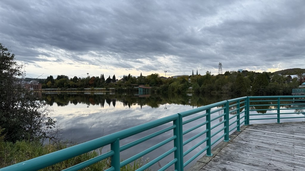 A tranquil riverside scene featuring a turquoise railing overlooking a calm waterway, with trees and buildings reflected in the water under a cloudy sky.