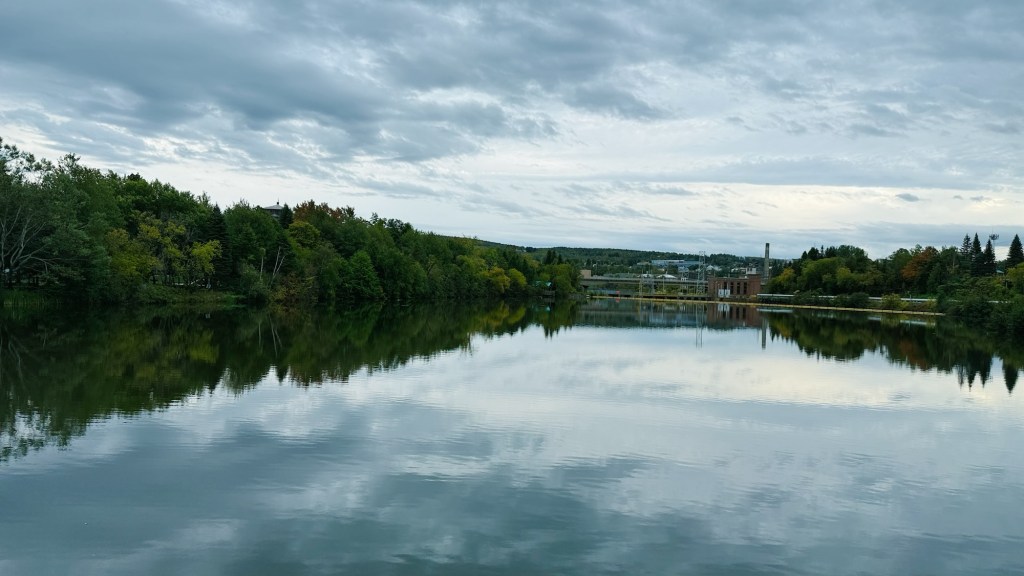 A tranquil lake reflecting a cloudy sky and surrounding trees, with a small building visible along the shore.