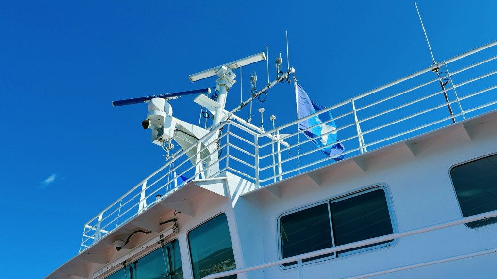 Close-up view of the top deck of a ship featuring navigation equipment, antennas, and a blue flag against a clear blue sky.