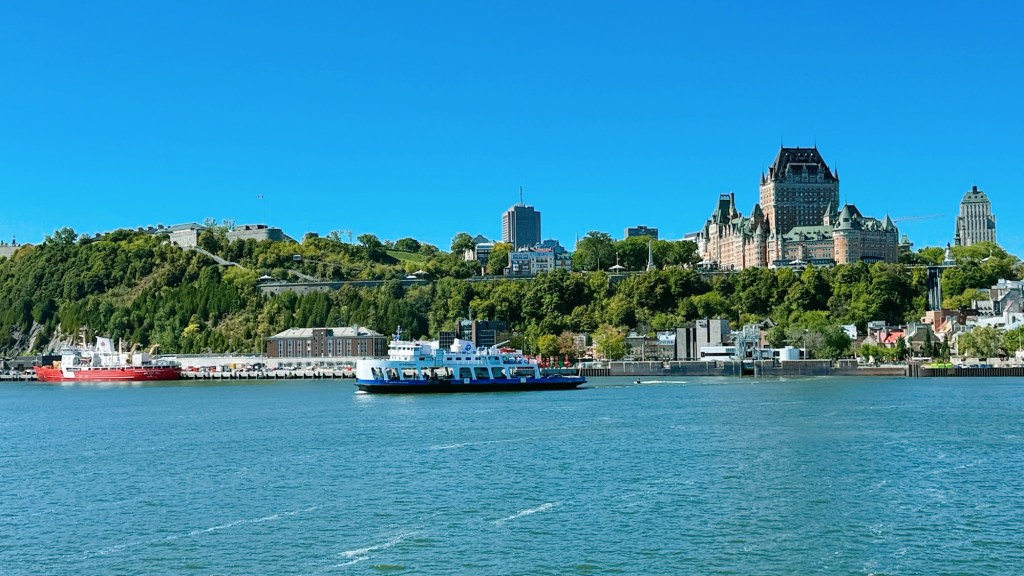 A scenic view of the Quebec City waterfront featuring a ferry sailing across the river, with lush greenery and historic buildings, including the iconic Château Frontenac, in the background against a clear blue sky.