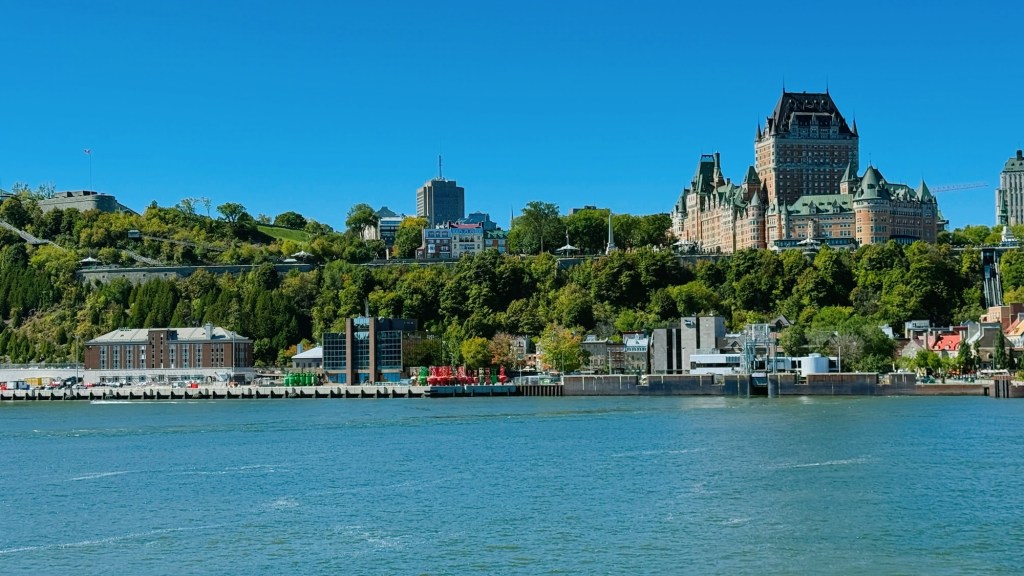 A view of the waterfront with green trees and buildings, featuring the iconic Château Frontenac in Quebec City against a clear blue sky.
