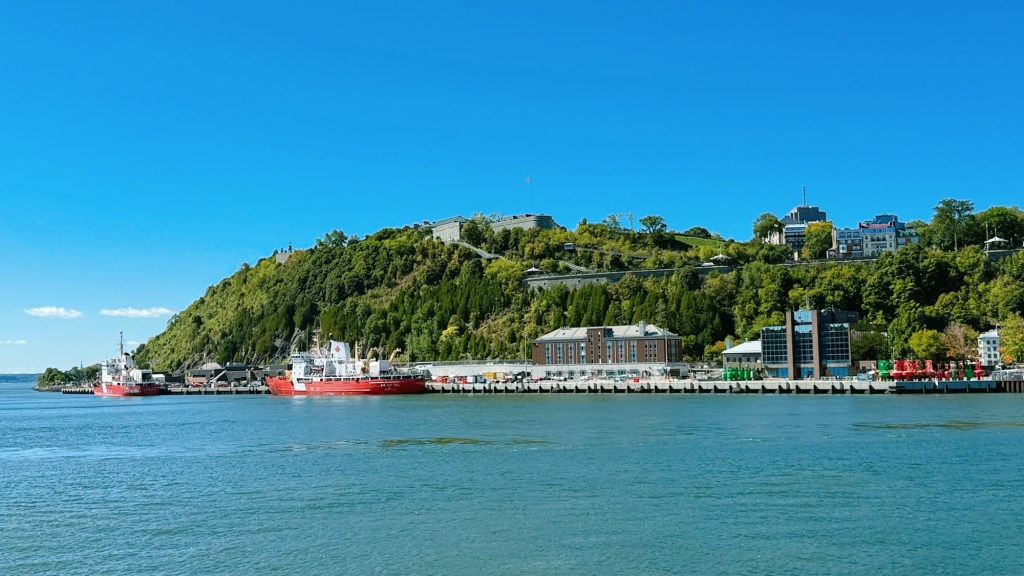 A panoramic view of a waterfront with red ships docked along a pier, surrounded by greenery and buildings on a hill against a clear blue sky.